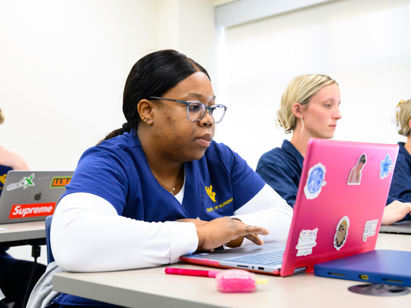 A nursing student in blue WVU School of Nursing scrubs works on a pink sticker‑covered laptop while others in blue scrubs use laptops in a classroom.