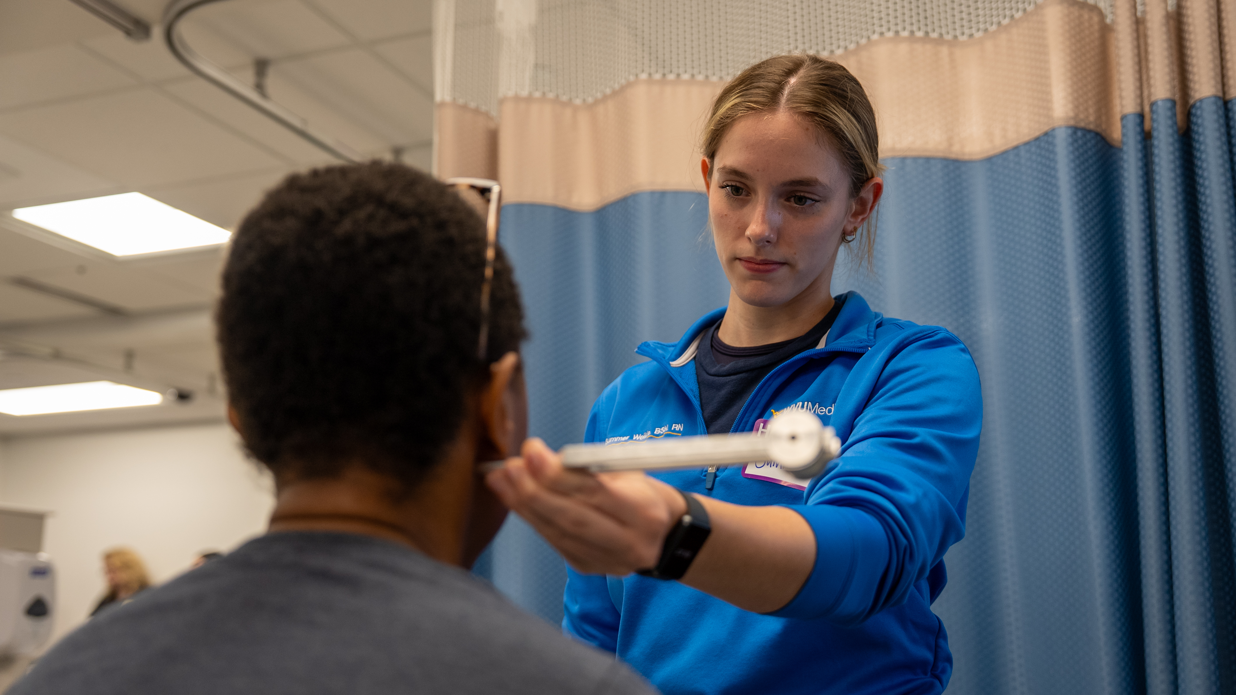 A graduate nursing student in blue scrubs stands beside and faces another person while using a handheld instrument to examine the area just below their ear in a medical setting.