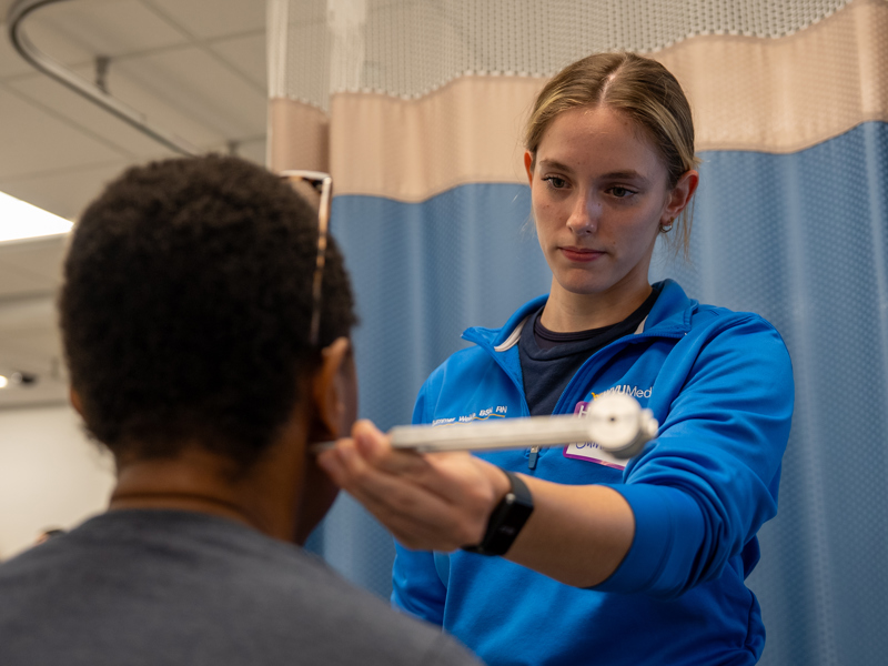 A graduate nursing student in blue scrubs stands beside and faces another person while using a handheld instrument to examine the area just below their ear in a medical setting.
