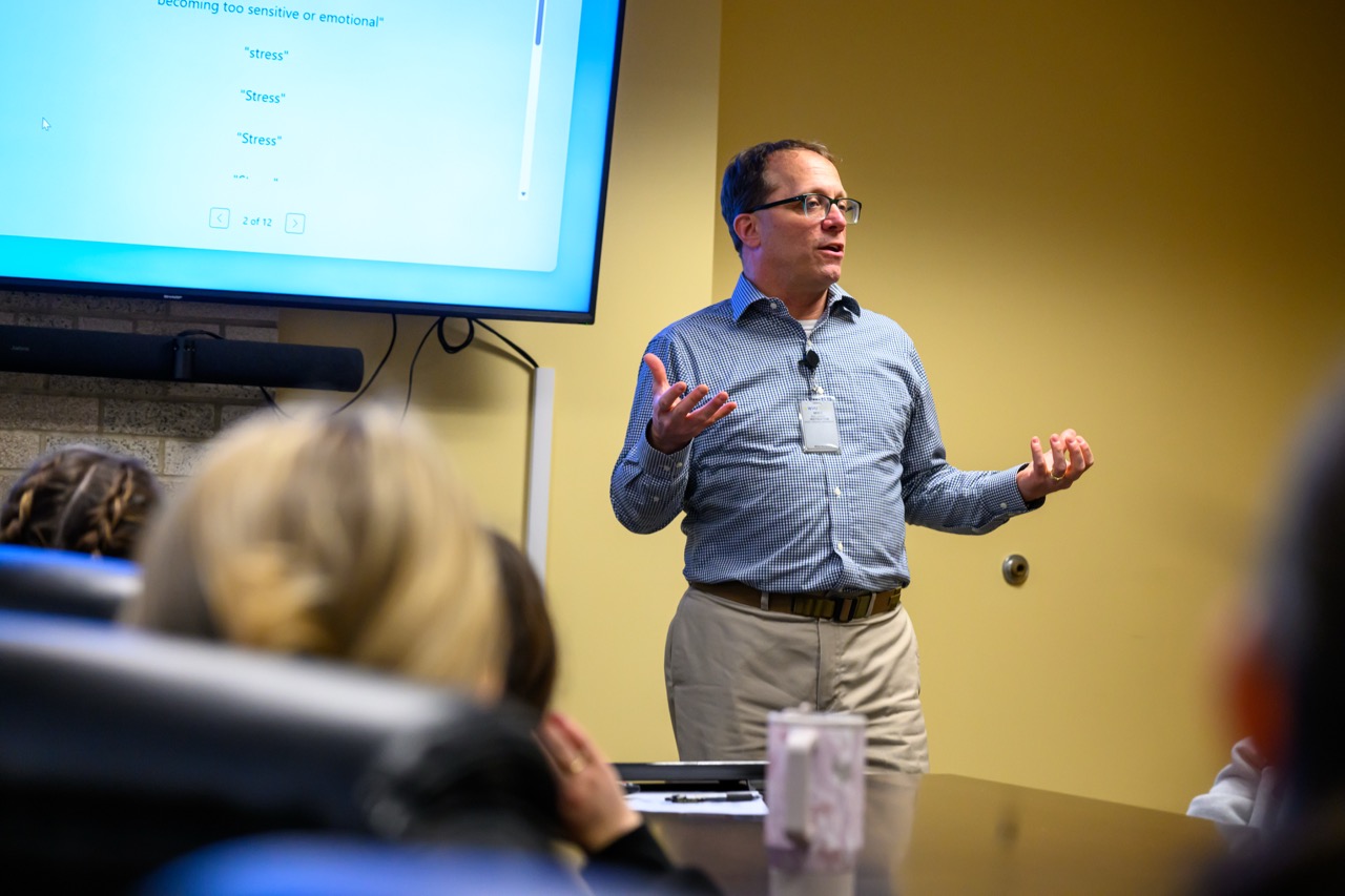 Presenter wearing glasses and a blue checkered shirt speaks to an audience while gesturing with both hands beside a presentation screen.