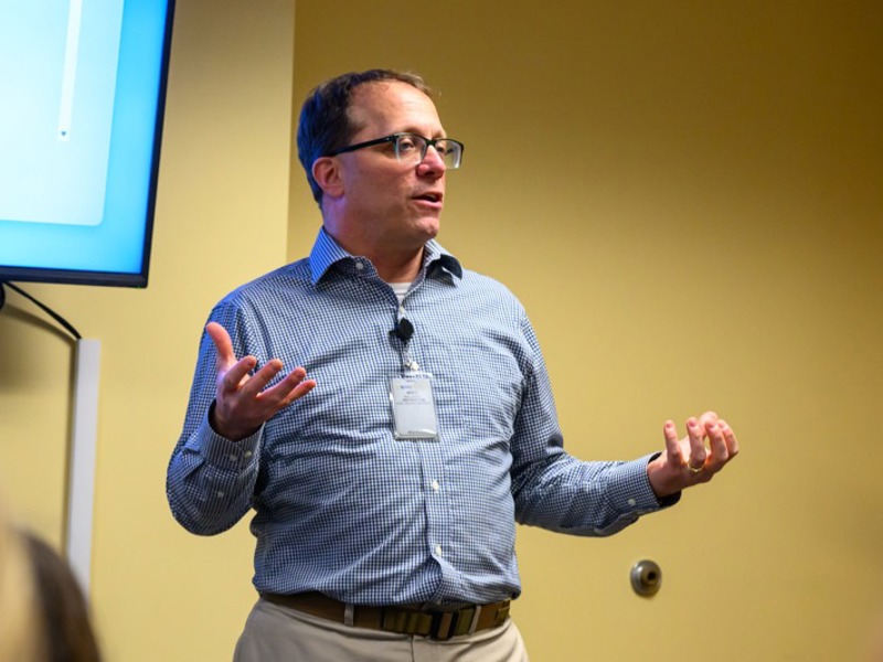 Presenter wearing glasses and a blue checkered shirt speaks to an audience while gesturing with both hands beside a presentation screen.