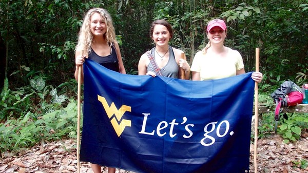 Three students hold up a West Virginia University “Let’s go.” flag and walking sticks while standing in a jungle-like setting.