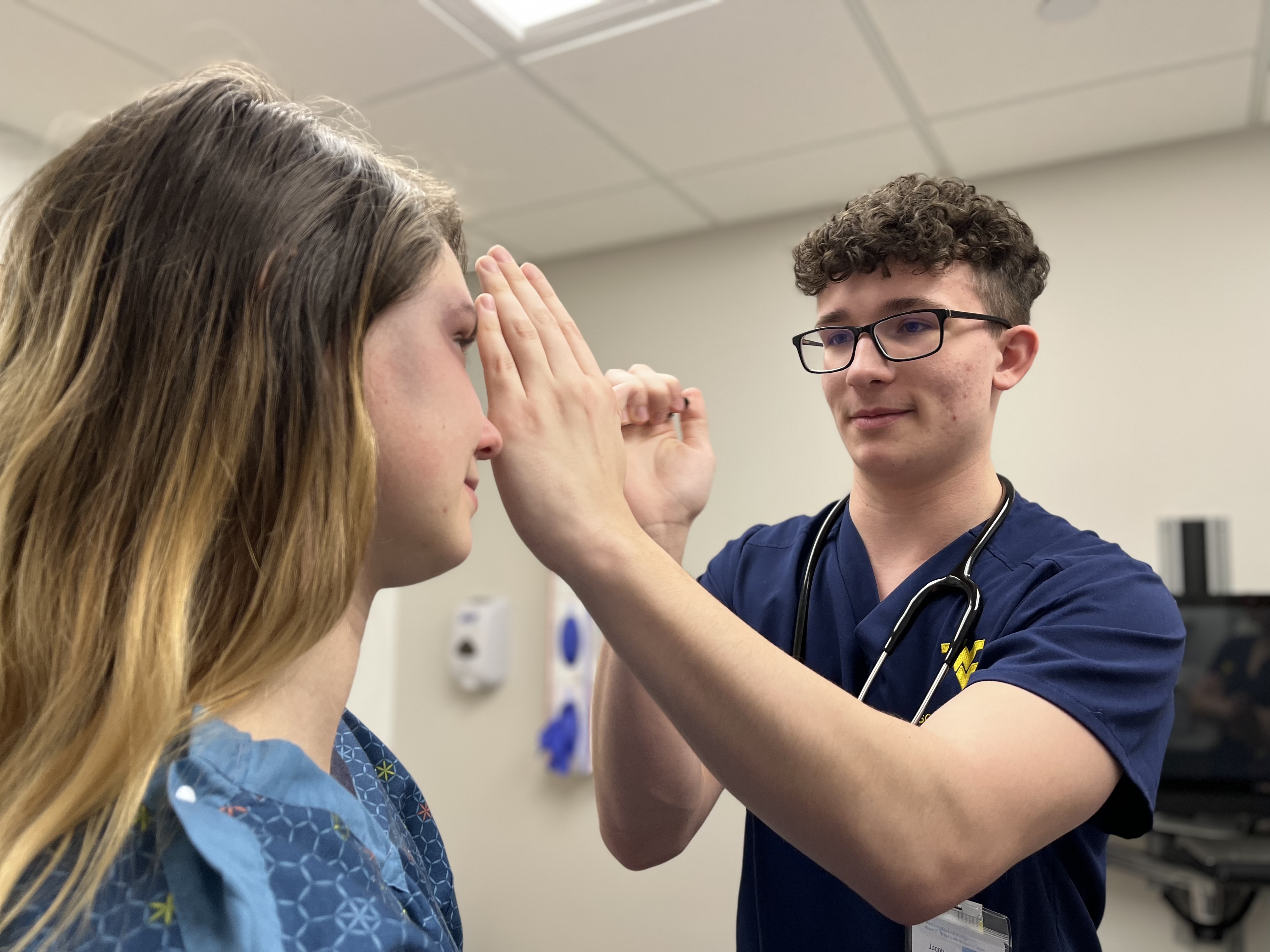 Male nursing student uses penlight to examine a patient in an exam room.