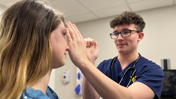 Male nursing student uses penlight to examine a patient in an exam room.