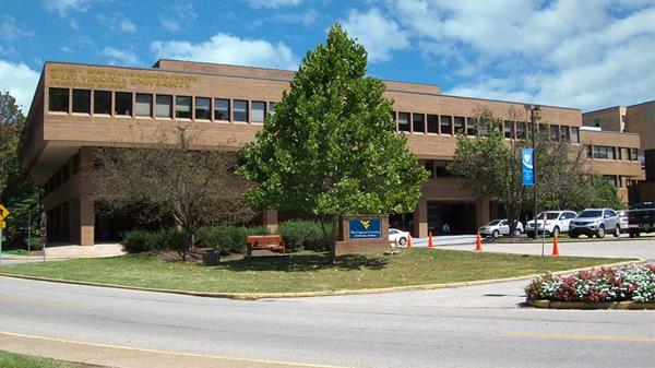 The exterior of the WVU Health Sciences Center Charleston Division building: a brick, brutalist-style building with one prominent tree out front. The building is beneath a blue sky with puffy white clouds. 