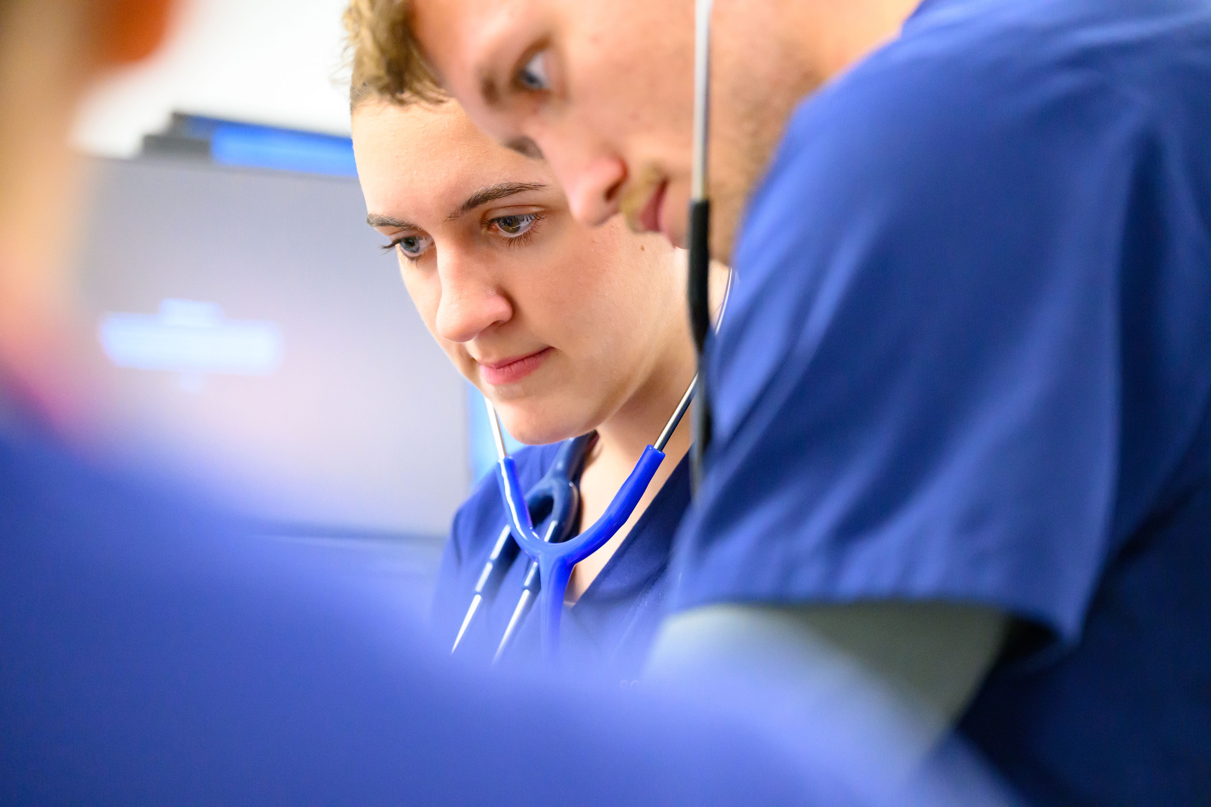 Two nursing students in blue scrubs with stethoscopes focus on a clinical task together in a skills lab.