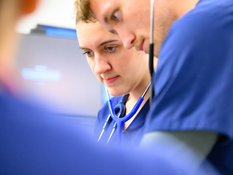 Two nursing students in blue scrubs with stethoscopes focus on a clinical task together in a skills lab.