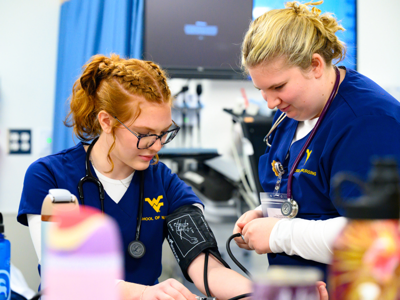A nursing student takes another student’s blood pressure in a clinical skills lab, with both wearing blue WVU School of Nursing scrubs.
