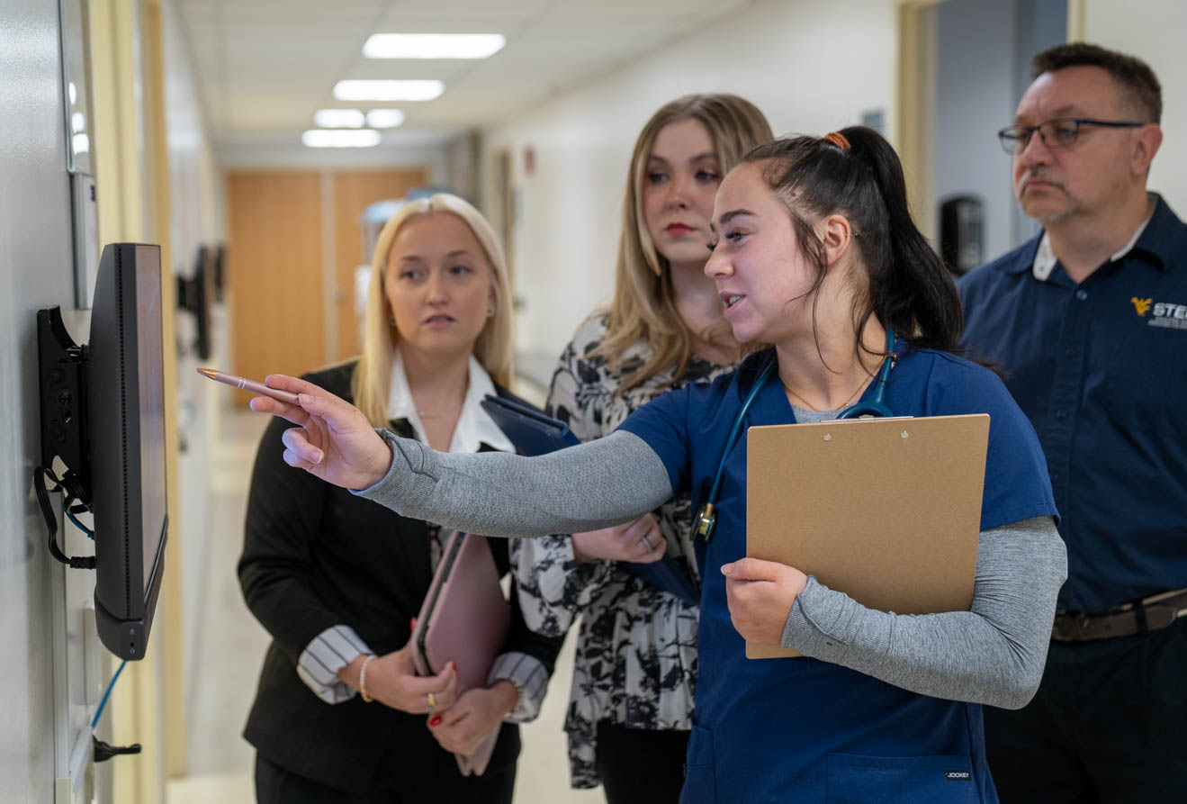A person in blue scrubs points to a wall‑mounted monitor while three colleagues observe in a hallway.