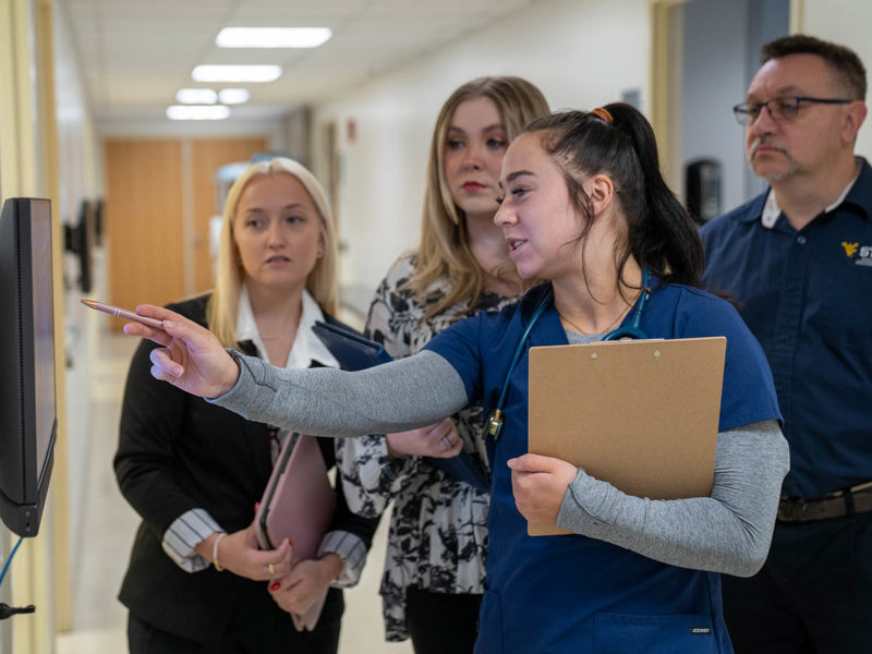 A person in blue scrubs points to a wall‑mounted monitor while three colleagues observe in a hallway.