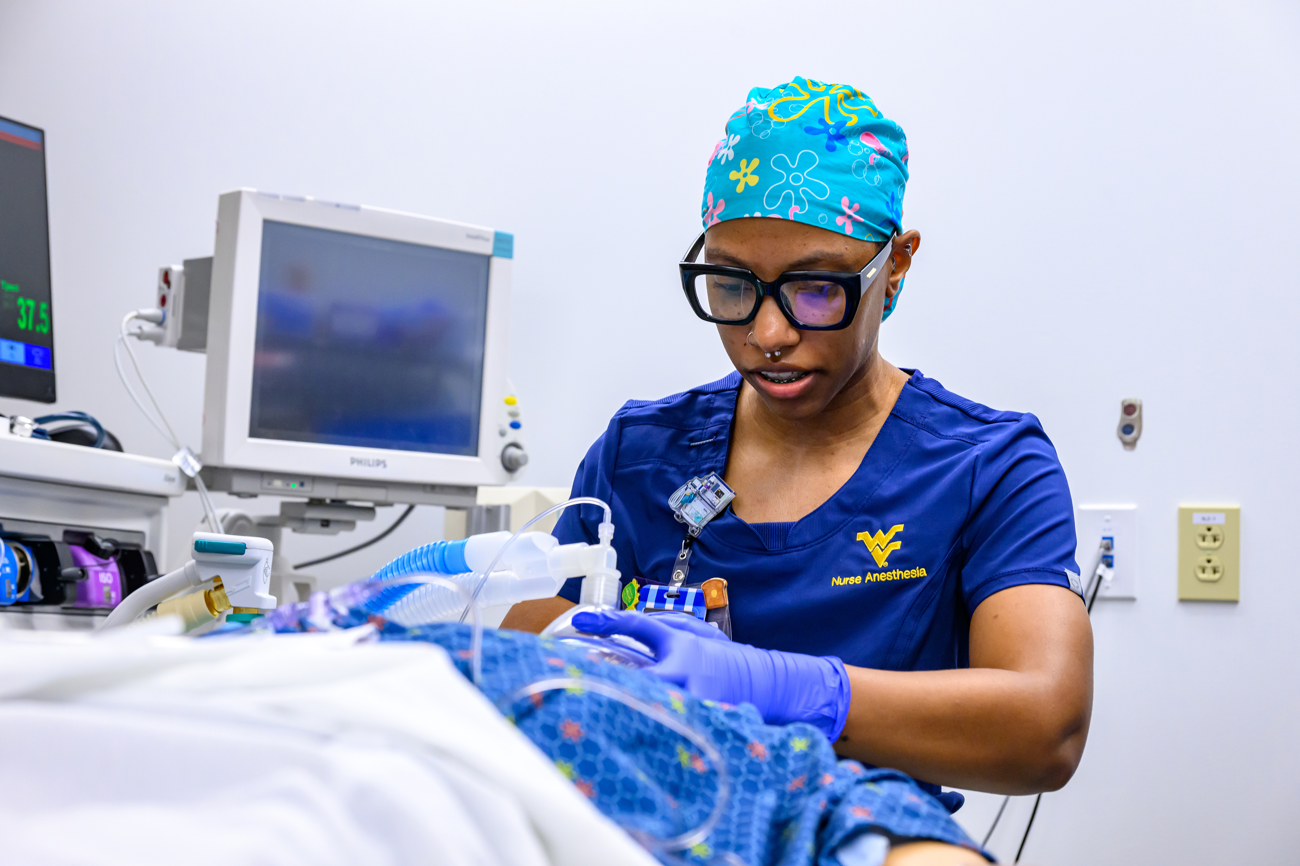 A nurse anesthetist in blue WVU Nurse Anesthesia scrubs and a colorful surgical cap adjusts anesthesia equipment beside a patient mannequin in a skills lab.