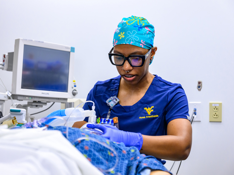 A nurse anesthetist in blue WVU Nurse Anesthesia scrubs and a colorful surgical cap adjusts anesthesia equipment beside a patient mannequin in a skills lab.