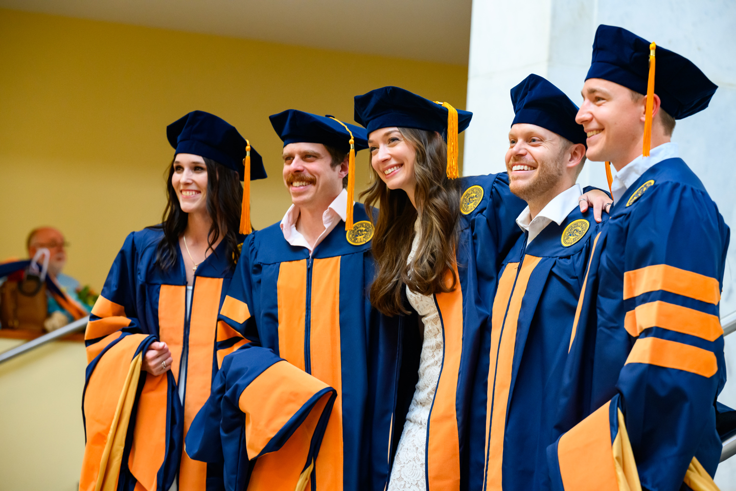 Five people in WVU blue academic gowns with gold panels and matching caps pose together smiling in front of a yellow wall at a commencement ceremony.