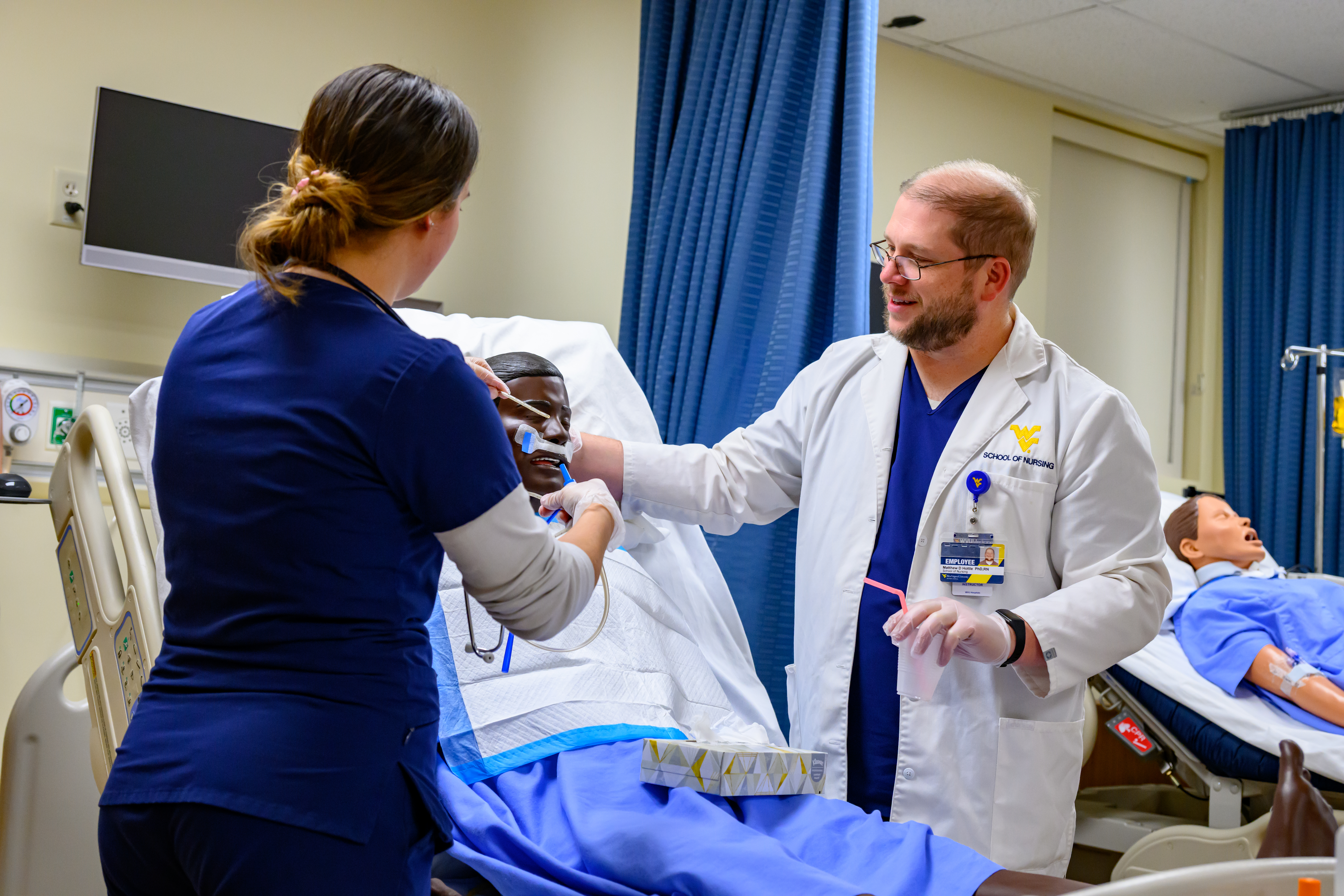 A student in blue scrubs adjusts tubing on a patient simulator while a faculty member in a white lab coat assists in a clinical training room with medical equipment and privacy curtains.