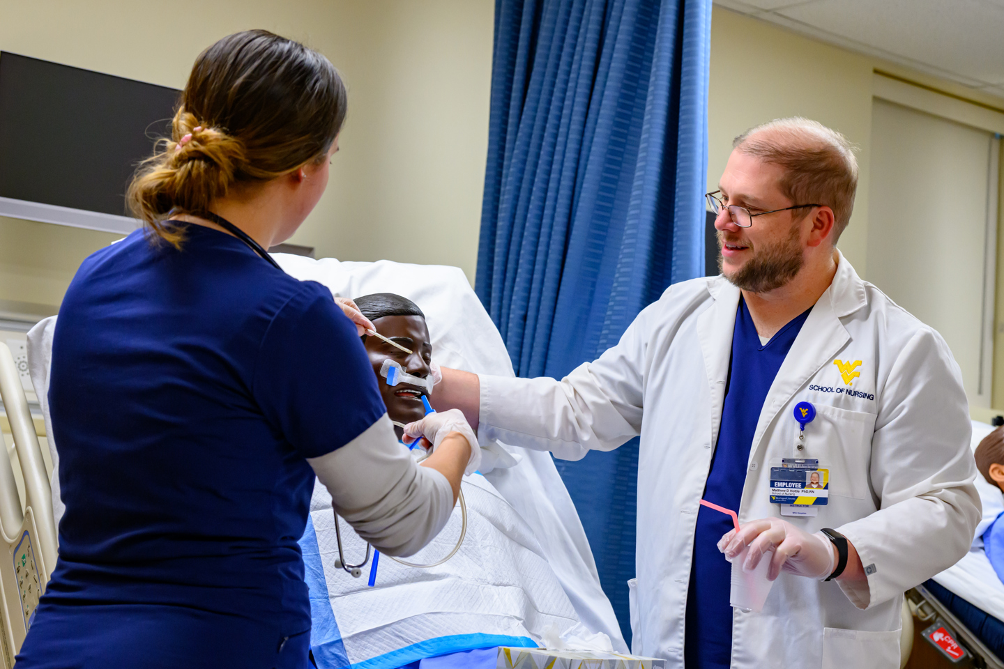 A student in blue scrubs adjusts tubing on a patient simulator while a faculty member in a white lab coat assists in a clinical training room with medical equipment and privacy curtains.