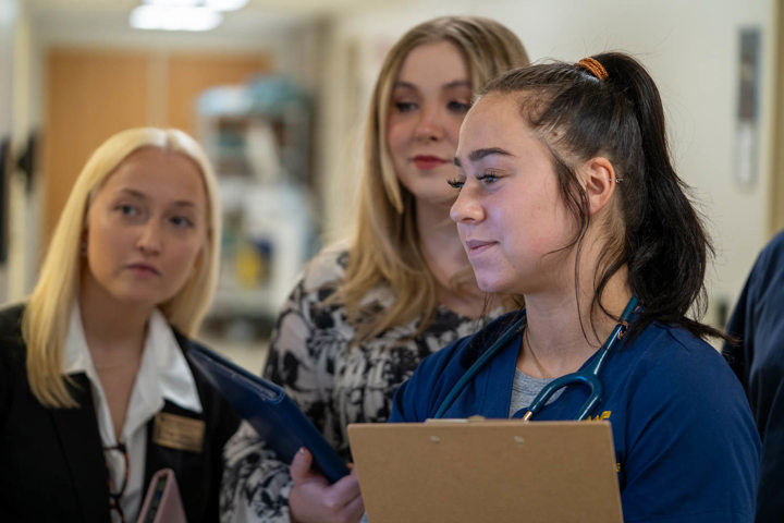 A nursing student in blue scrubs with a stethoscope holds a clipboard while speaking with two professionally dressed individuals in a hallway.