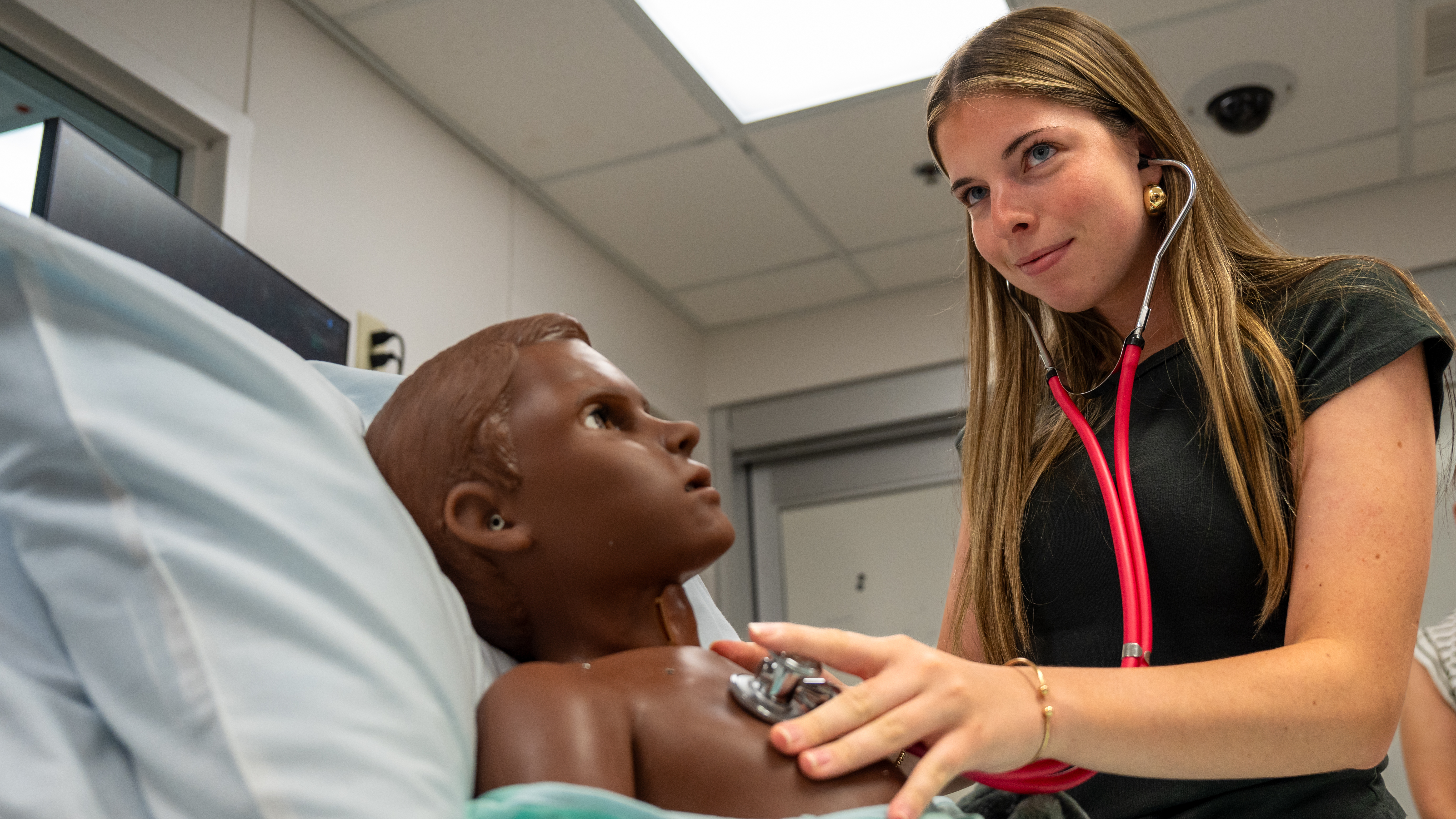 A student in black scrubs holds a stethoscope to the chest of a manikin.