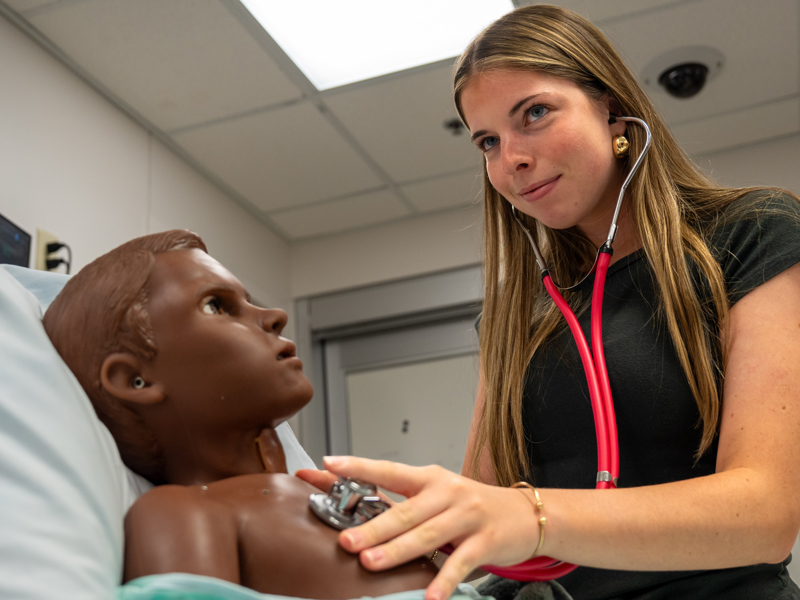 A student in black scrubs holds a stethoscope to the chest of a manikin.