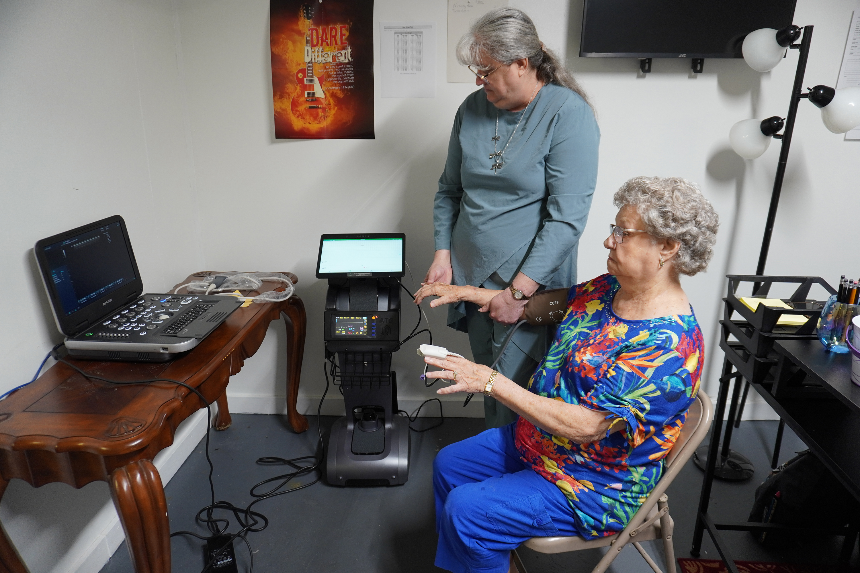 An elderly woman sits with a blood pressure cuff and finger sensor while a clinician stands beside her operating diagnostic equipment in a small office‑like room.