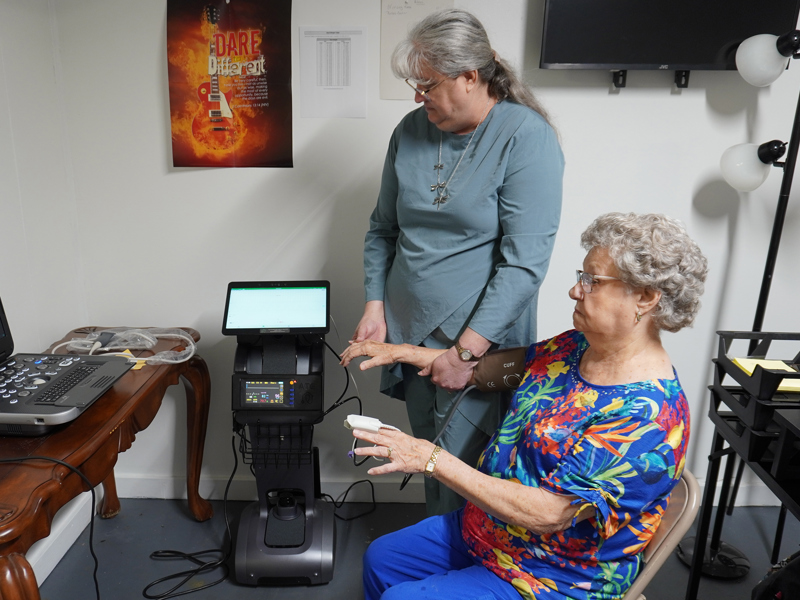 An elderly woman sits with a blood pressure cuff and finger sensor while a clinician stands beside her operating diagnostic equipment in a small office‑like room.