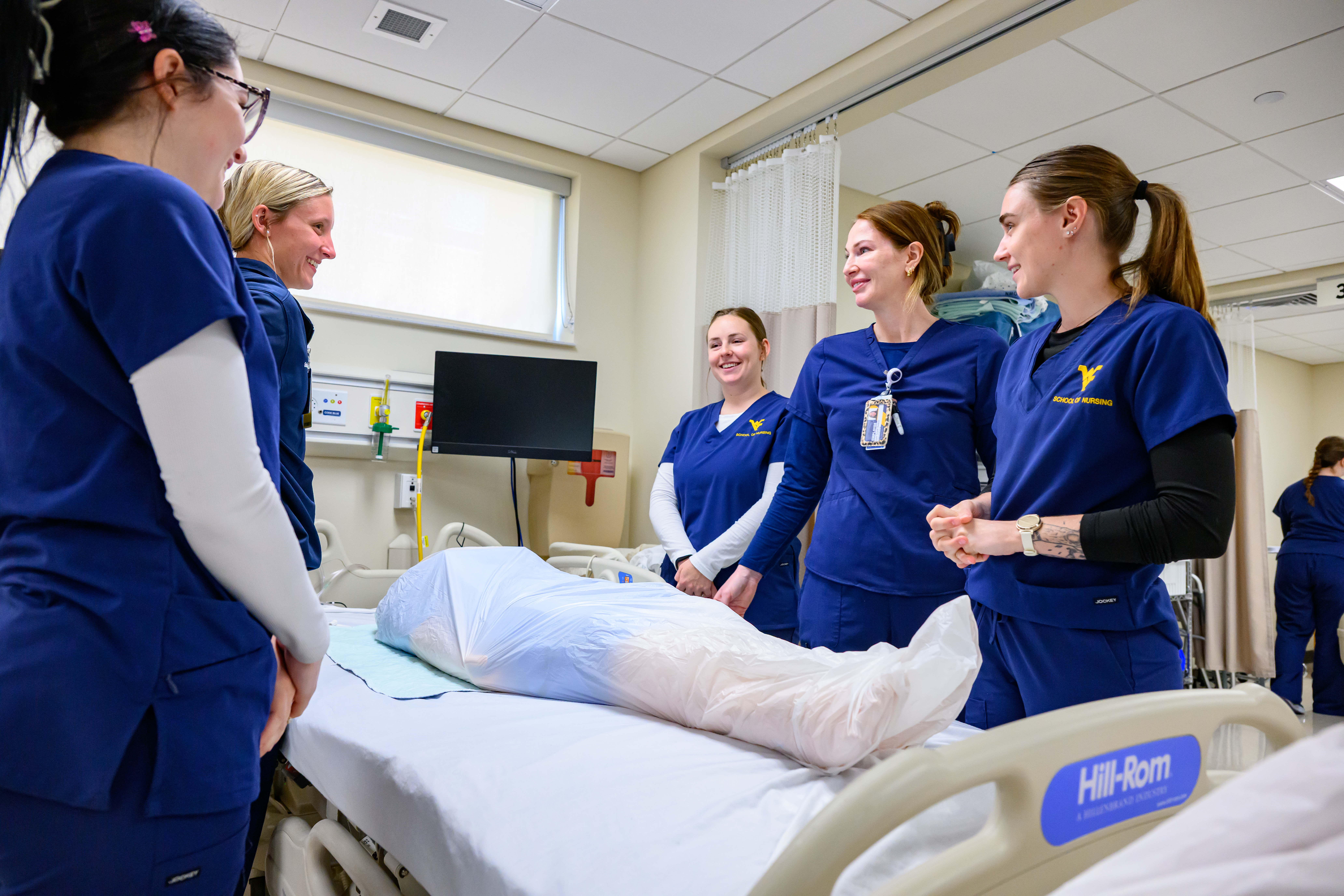 Three nursing students in blue WVU School of Nursing scrubs stand together in a clinical skills lab, smiling in front of a privacy curtain.
