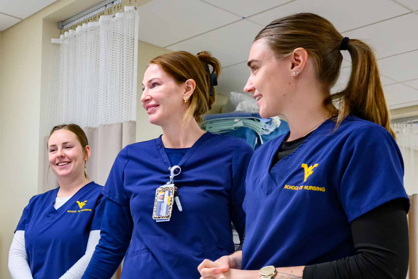 Three nursing students in blue WVU School of Nursing scrubs stand together in a clinical skills lab, smiling in front of a privacy curtain.