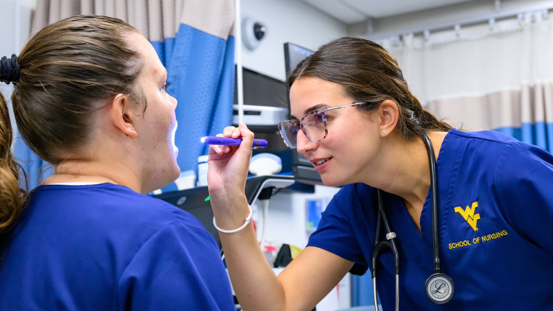 A student in blue scrubs uses a penlight to examine another student’s open mouth in a clinical training room with medical equipment and privacy curtains.
