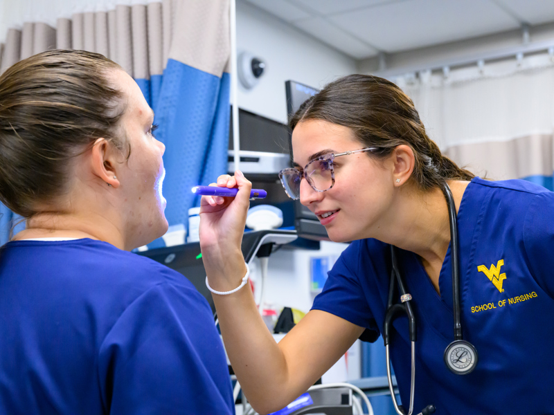 A student in blue scrubs uses a penlight to examine another student’s open mouth in a clinical training room with medical equipment and privacy curtains.