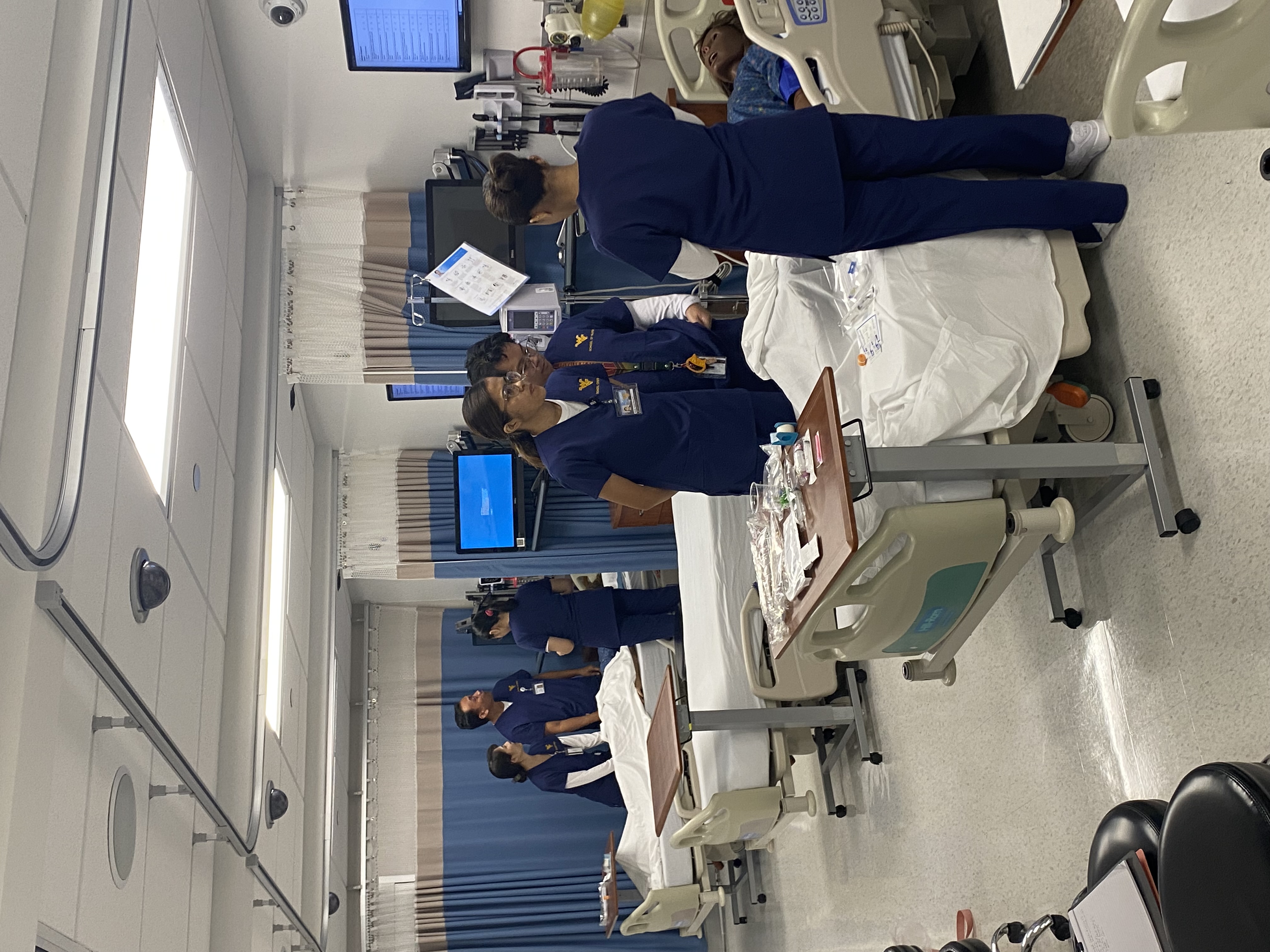 Three Filipino nursing students in blue WVU School of Nursing scrubs stand around a hospital bed with a simulation mannequin in a clinical skills lab.