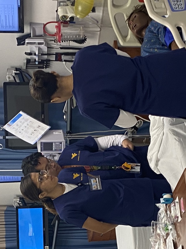 Three Filipino nursing students in blue WVU School of Nursing scrubs stand around a hospital bed with a simulation mannequin in a clinical skills lab.