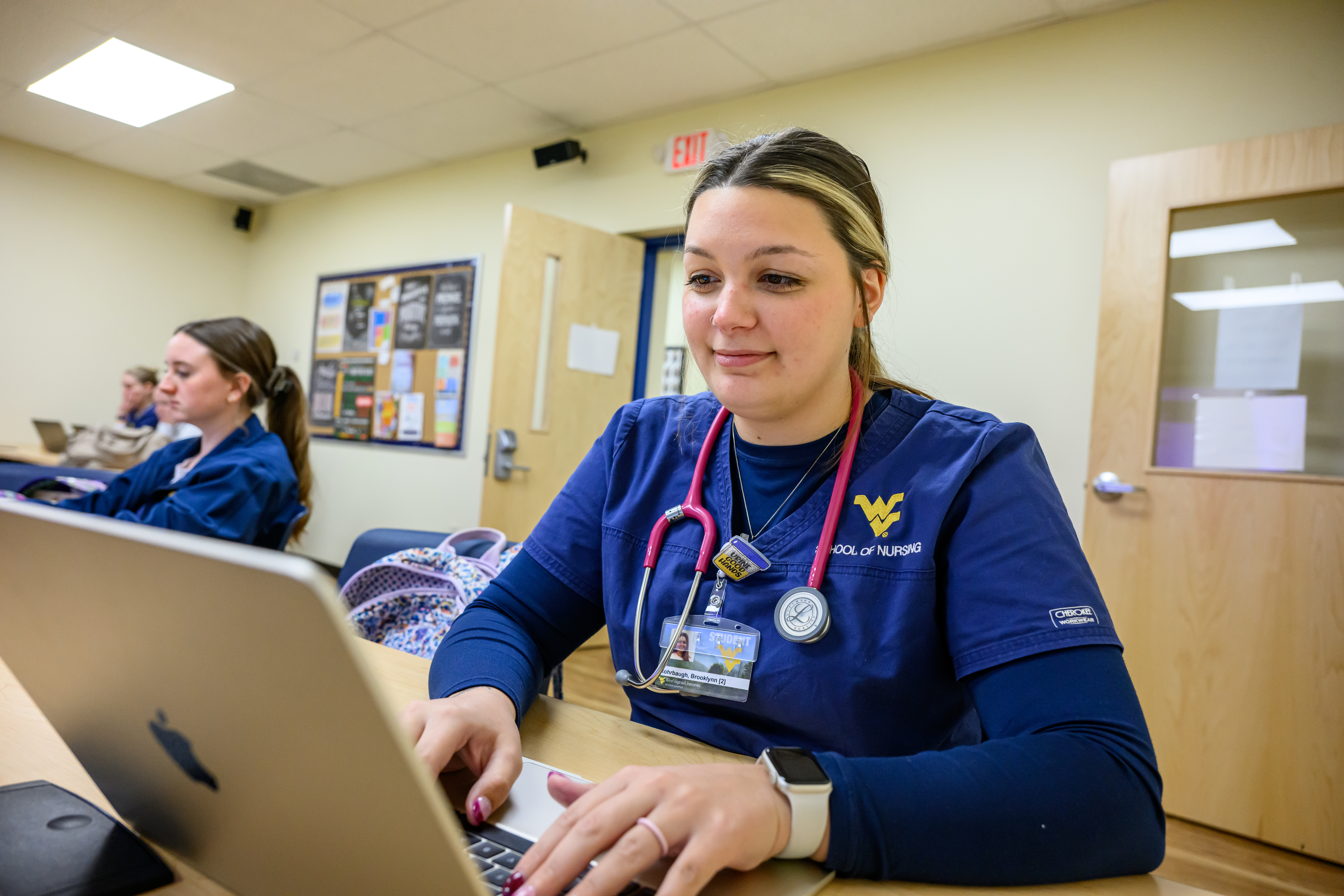 A nursing student in blue WVU School of Nursing scrubs works on a laptop at a desk while another student in similar attire sits behind them in a classroom.