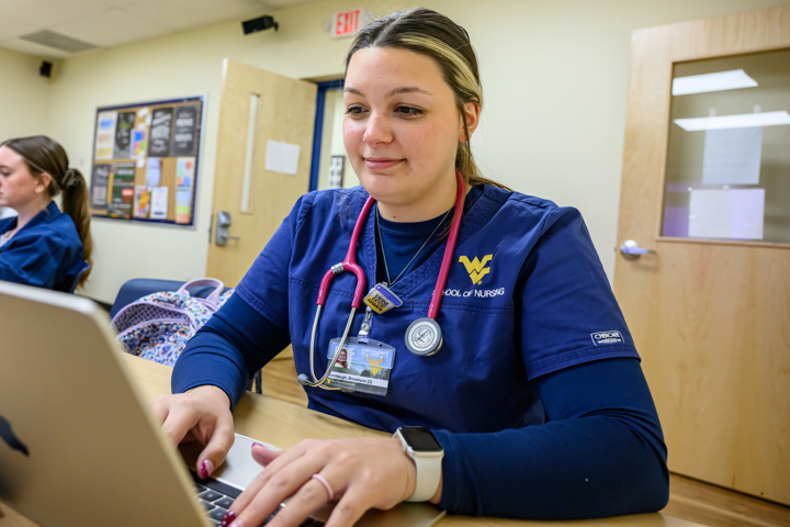 A nursing student in blue WVU School of Nursing scrubs works on a laptop at a desk while another student in similar attire sits behind them in a classroom.