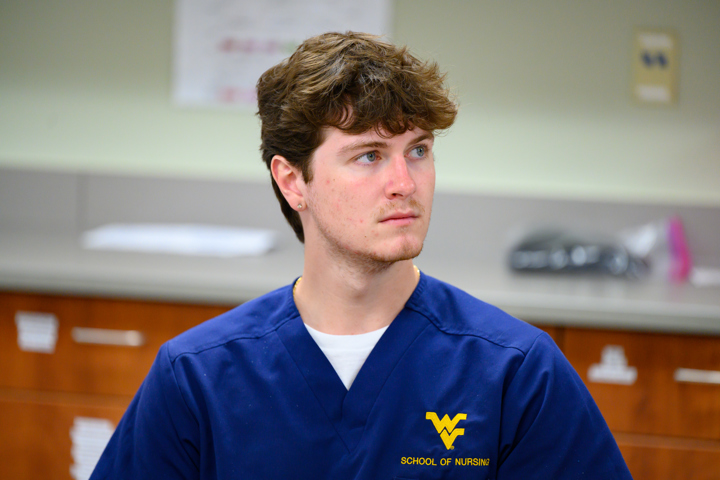 A nursing student in blue WVU School of Nursing scrubs sits in a classroom or lab with cabinets in the background.