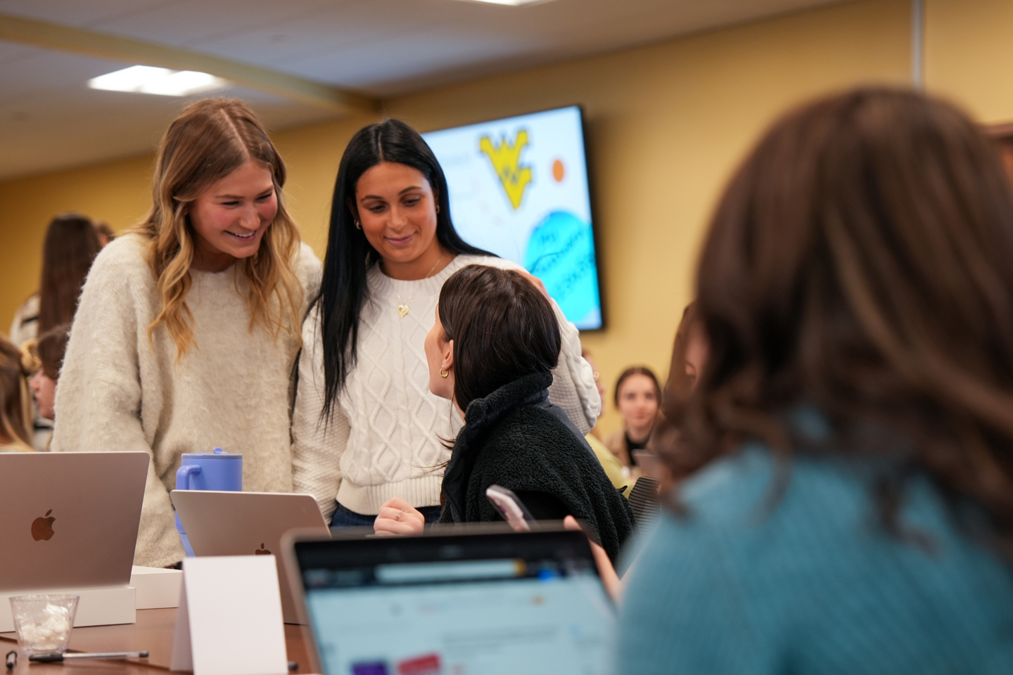 Three students interact around a table with laptops and notebooks, with two standing and talking to one seated, in a classroom full of students.