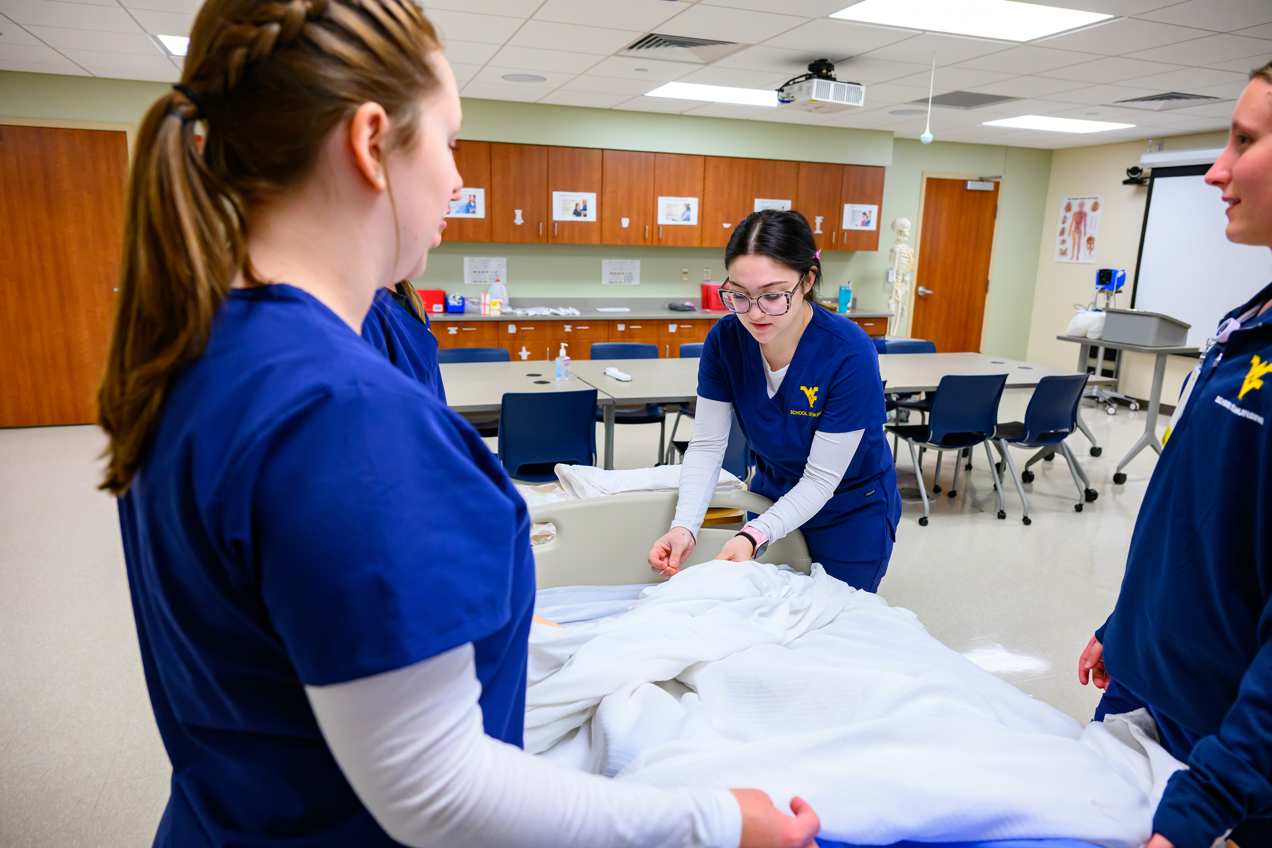 One nursing student adjusts linens on a hospital bed while two others observe in a skills lab with classroom equipment in the background.