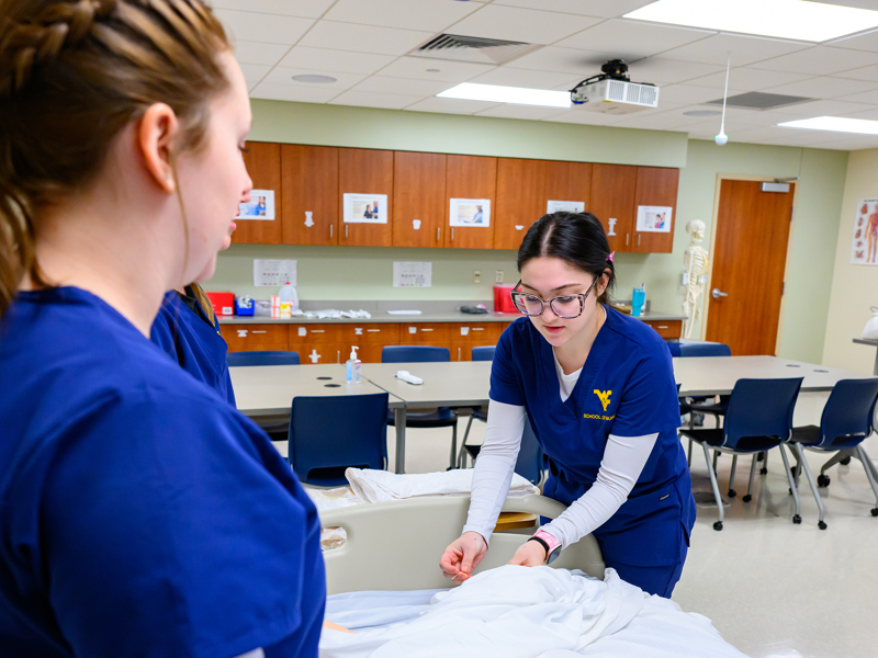 One nursing student adjusts linens on a hospital bed while two others observe in a skills lab with classroom equipment in the background.