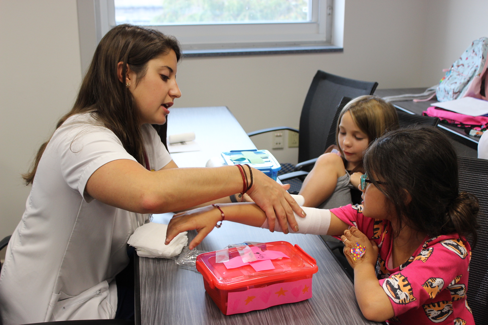 A nurse, sitting to the left side of a table, bandages the arm of a young girl sitting on the opposite side of the table. In the background, another young girl sits and watches.