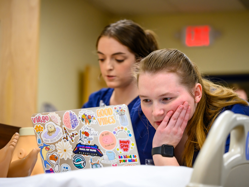 Two students in blue scrubs look at a sticker‑covered laptop beside a medical training mannequin in a simulation lab.