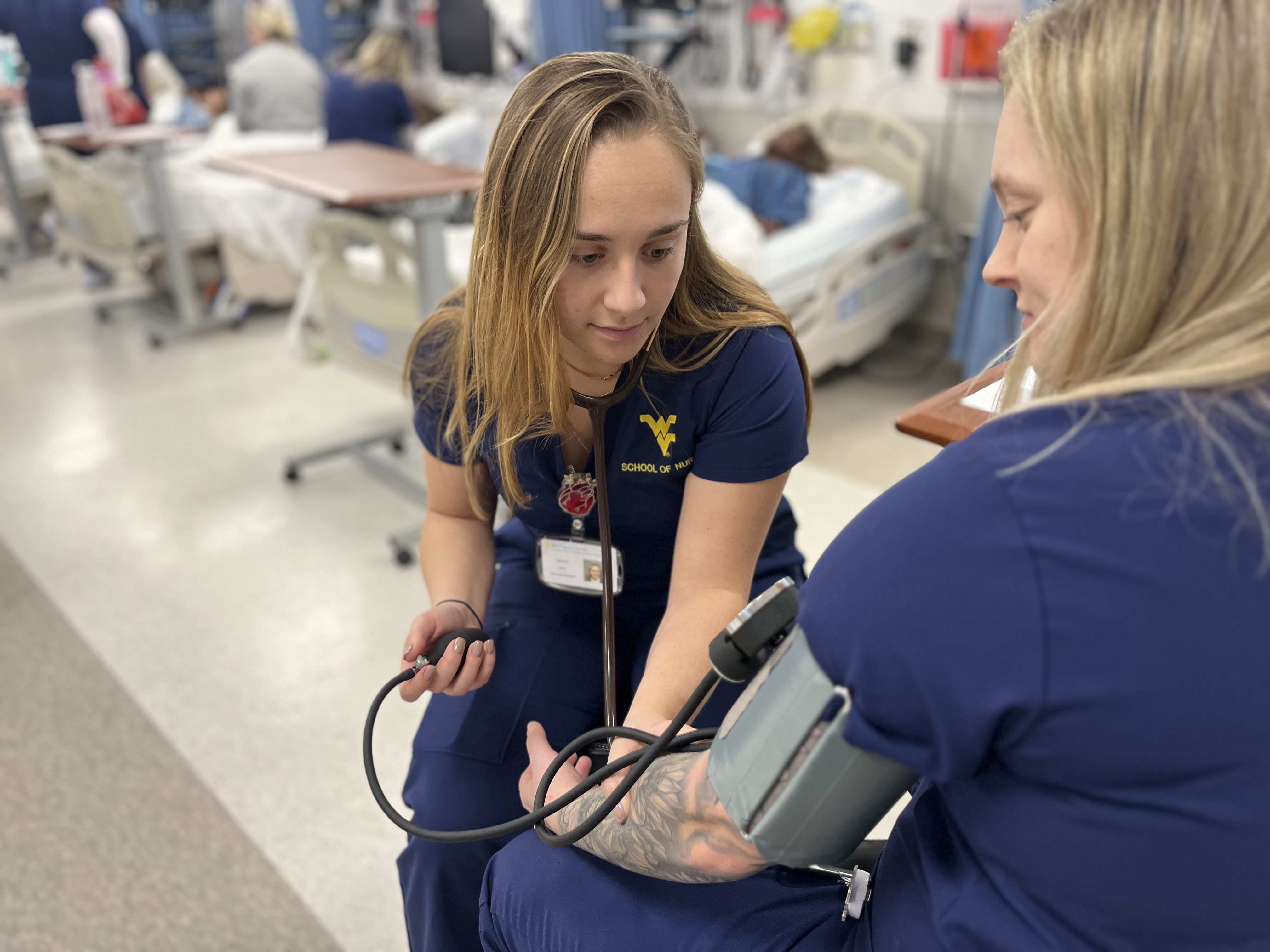 A student wearing blue scrubs checks the blood pressure of another student using an arm cuff. 