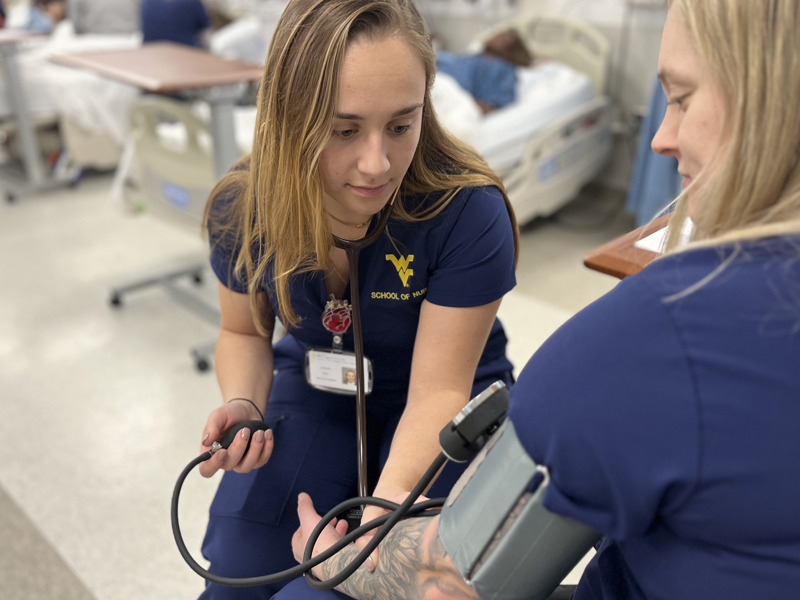 A student wearing blue scrubs checks the blood pressure of another student using an arm cuff. 