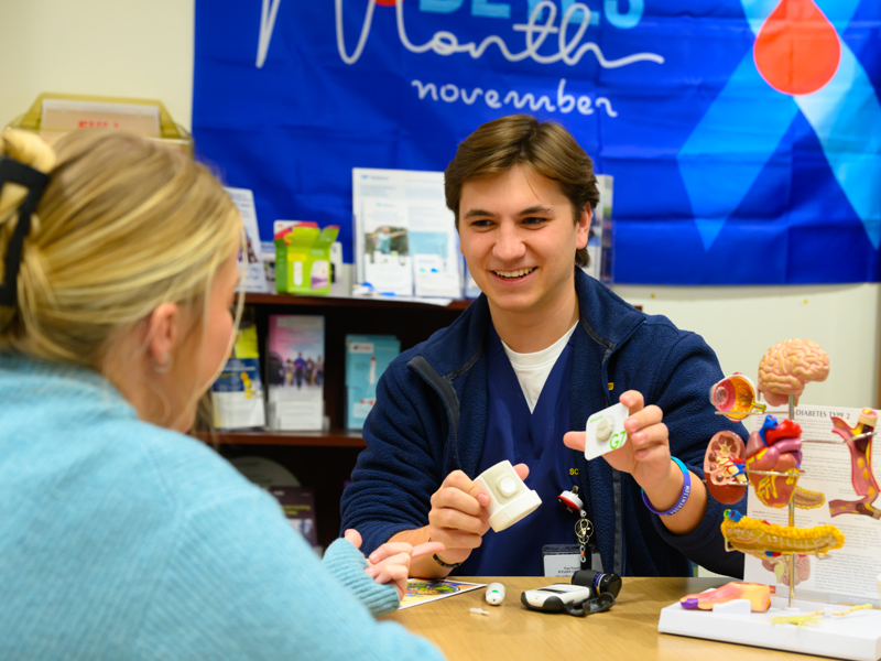 A student wearing blue scrubs and a navy jacket shows medical devices and anatomical models to another person at a table, with a “Diabetes Month November” banner on the wall behind them.