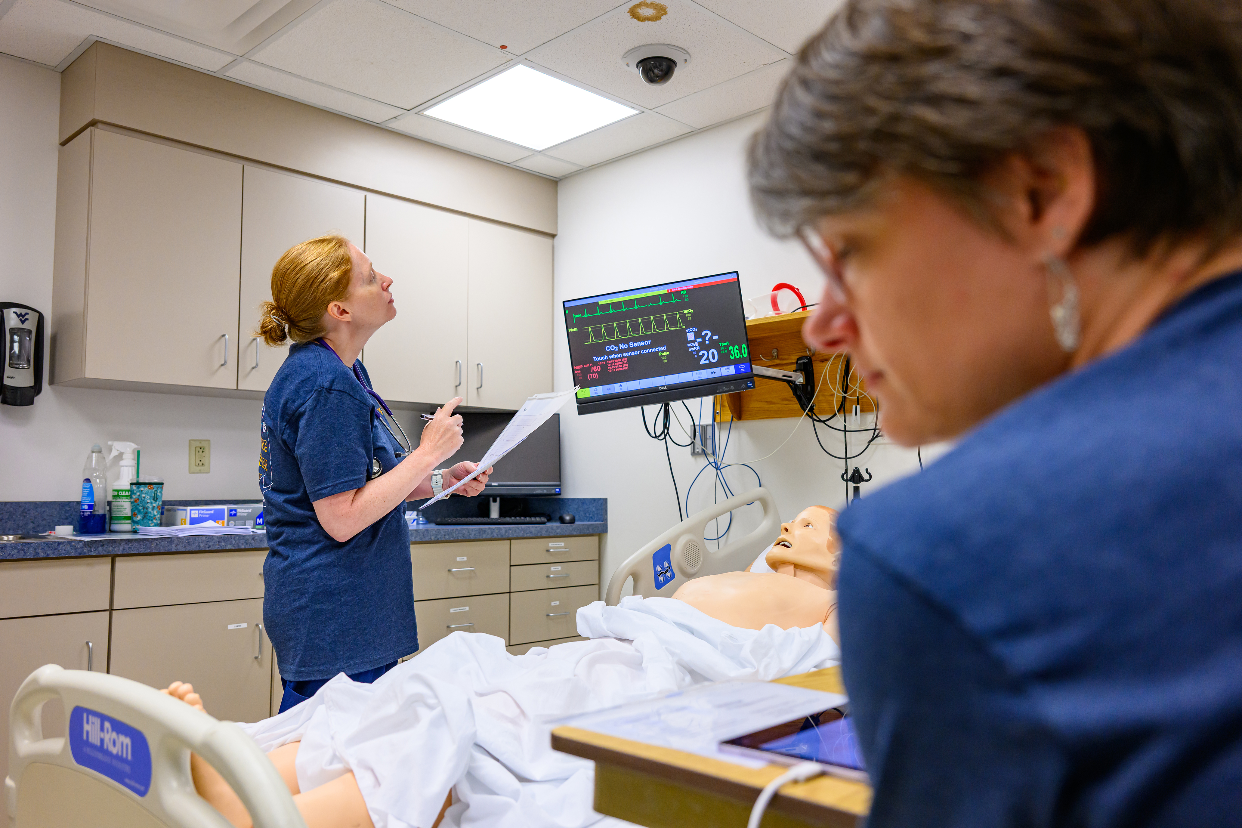 A person stands beside a hospital bed holding papers and looking at a patient monitor. A training mannequin lies in the bed, and another person is seated in the foreground.
