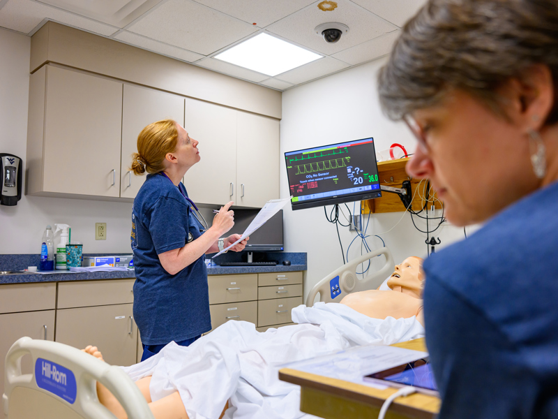 A person stands beside a hospital bed holding papers and looking at a patient monitor. A training mannequin lies in the bed, and another person is seated in the foreground.