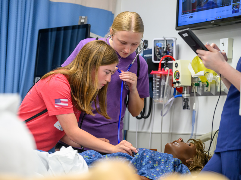 Two nursing students use a stethoscope to assess a simulation mannequin while another records the exercise on a tablet in a clinical training room.
