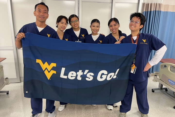 Six students stand together holding a large blue Let’s Go! flag in a clinical training room.