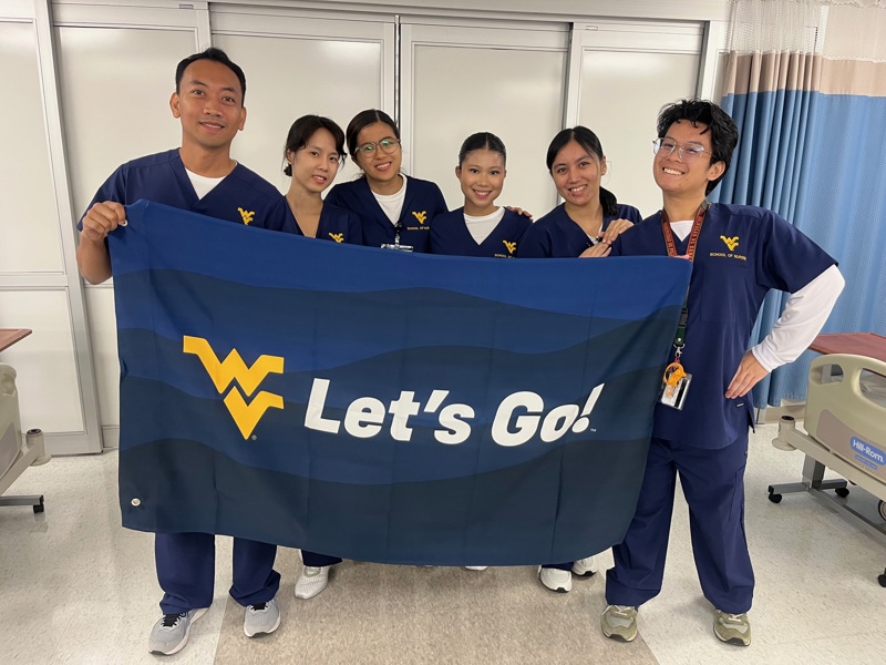 Six students stand together holding a large blue Let’s Go! flag in a clinical training room.
